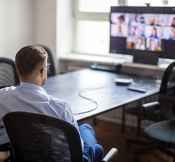 Business man viewed from behind at small conference table with Microsoft Teams video conference on large monitor.