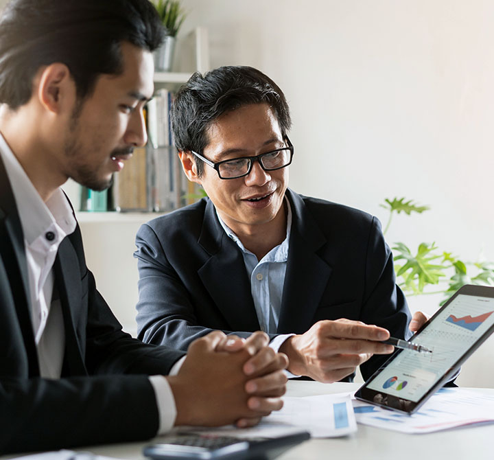 Two male business professionals, sitting at a table, are working on a presentation using a tablet device.