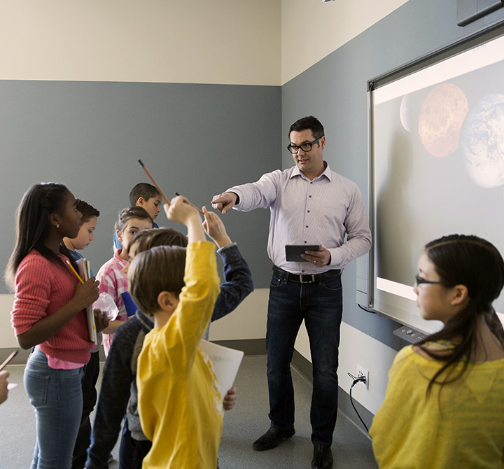 A teacher, holding a mobile device, is conducting a class with students all standing in front of the classroom.