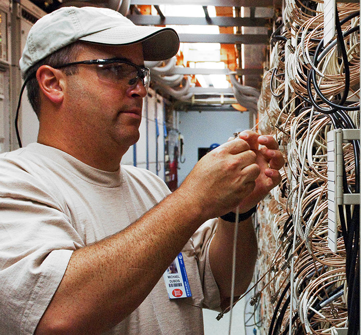 A technician wearing a beige t-shirt, hat and protective eye glasses, is working on wiring in a data room.