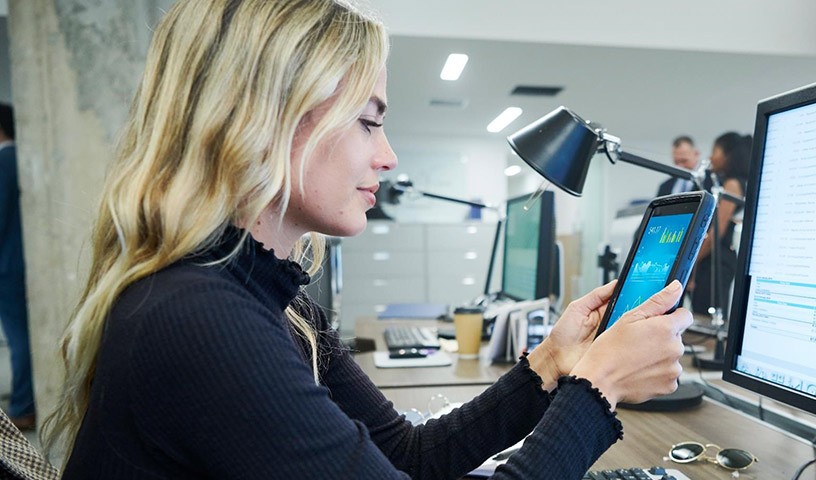 A woman is sitting in front of a computer monitor and keyboard while holding a wireless device she is using for work.