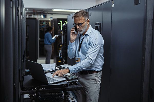 Male IT technician using a laptop while talking on a phone in a server room